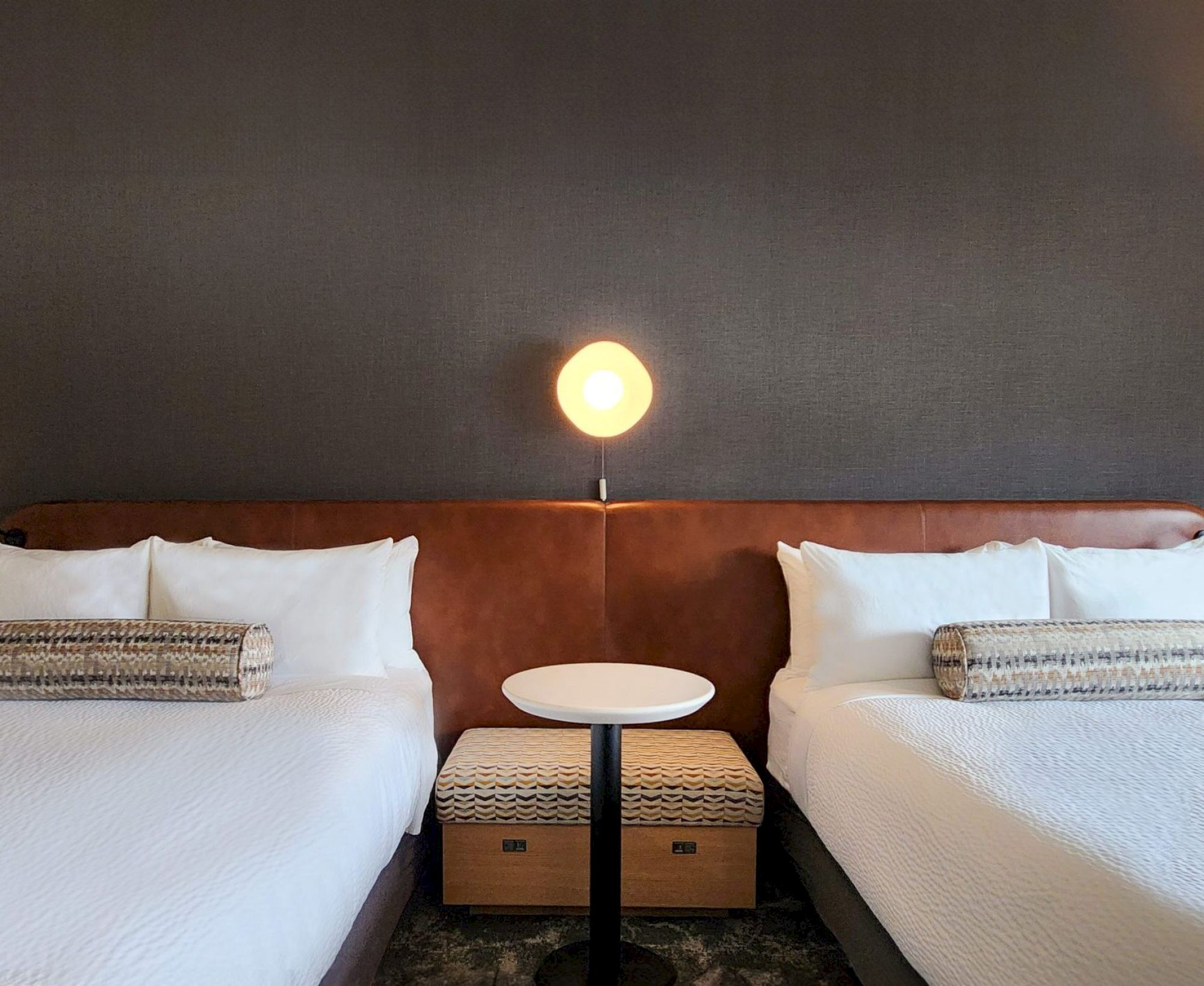 The image shows a hotel room with two beds, a round table in between, and a lamp above it on a dark wall.