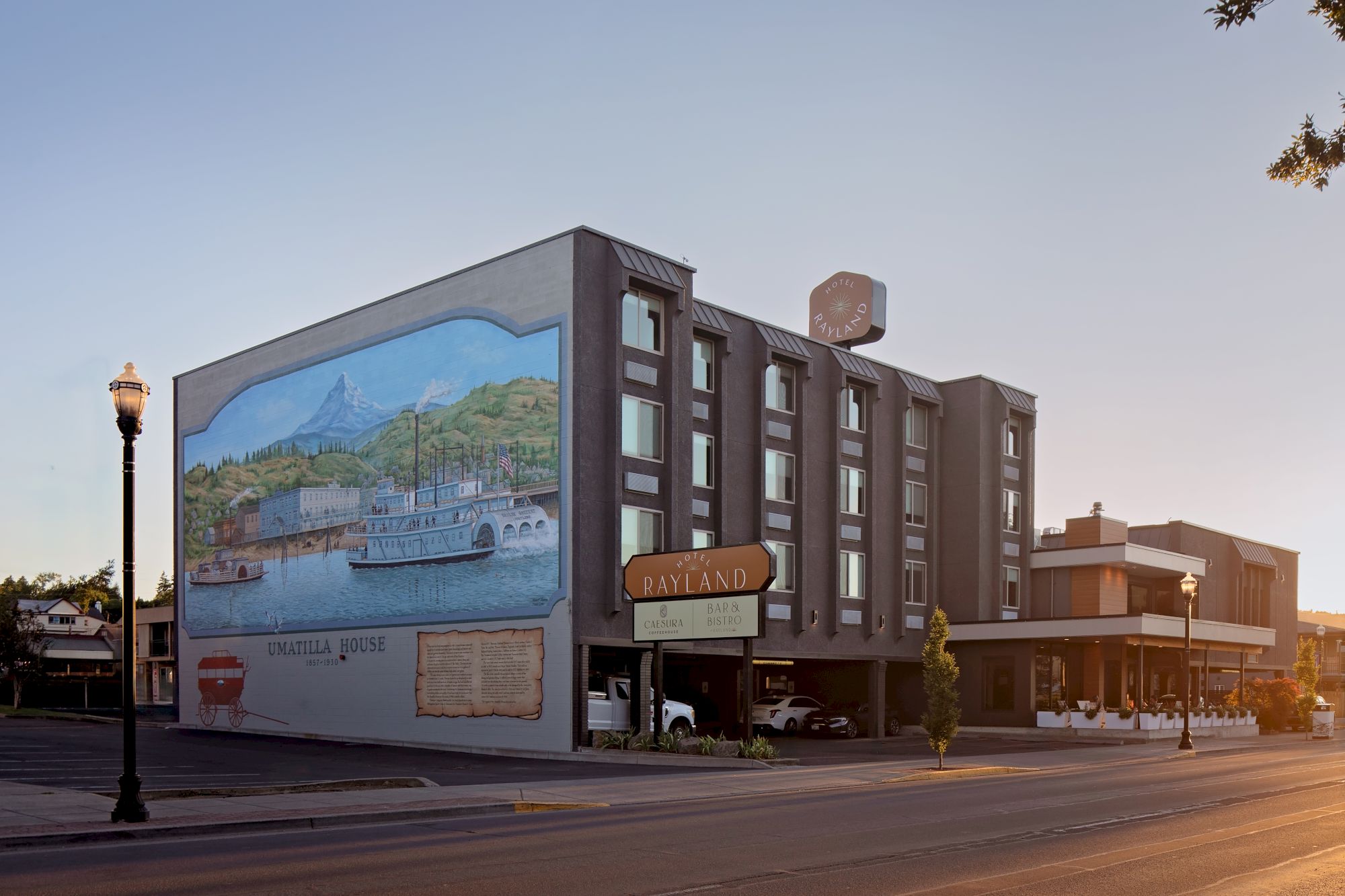 A brown hotel building labeled Hotel Rayland with cafe/bar signs and a mural showing a riverboat and mountains.