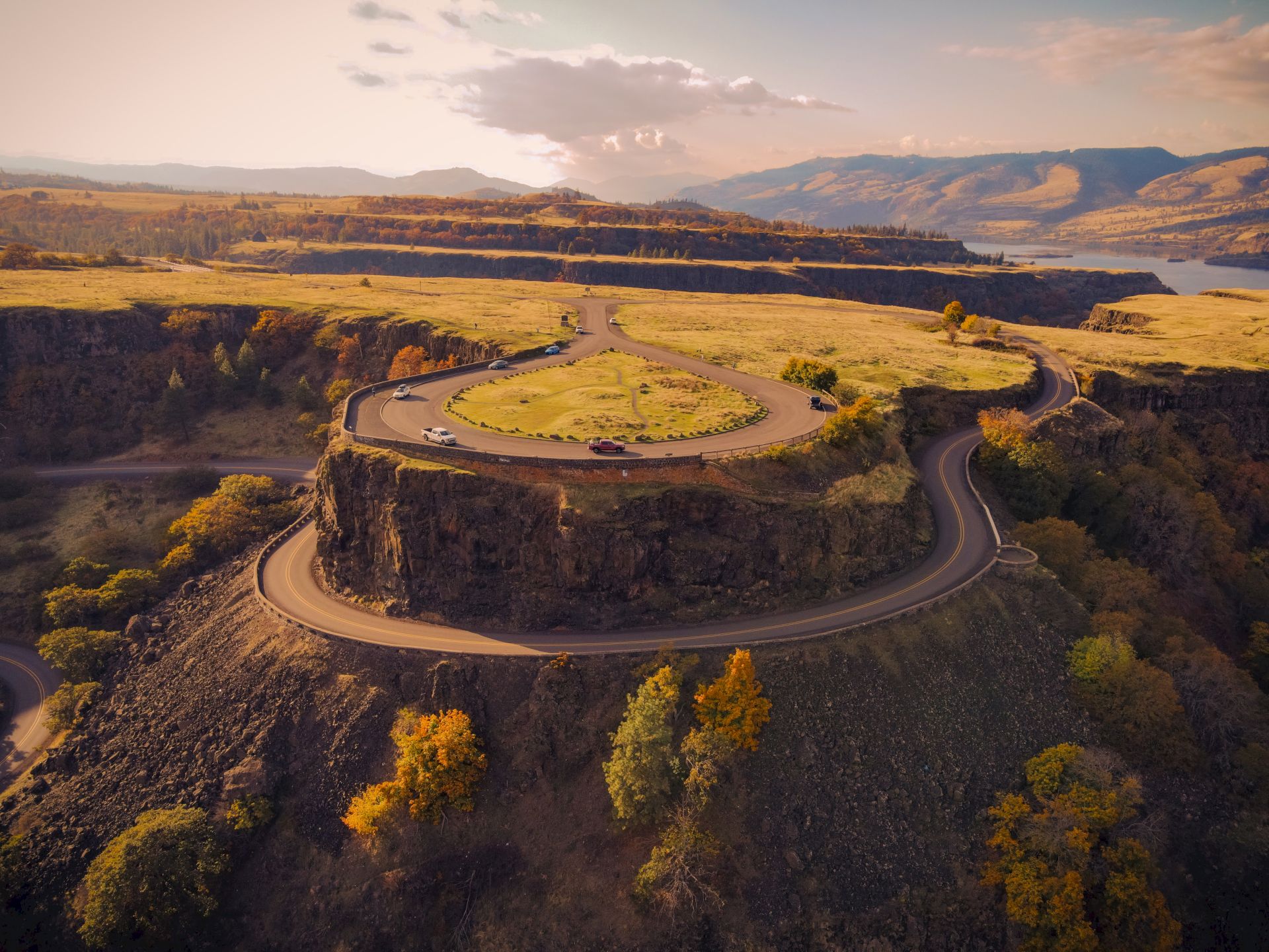 A winding road leads to a circular viewpoint atop a cliff, surrounded by autumn foliage with mountains and a river in the background.