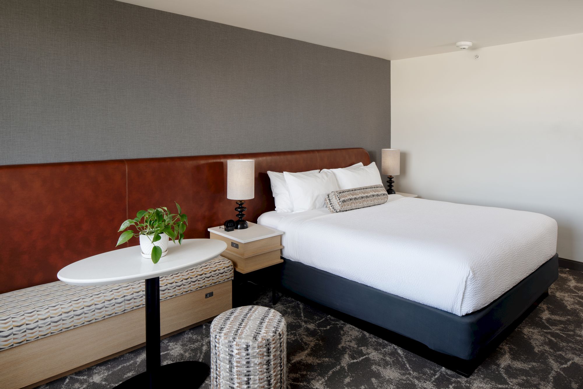 A modern hotel bedroom with a large bed, white linens, nightstands, lamps, and a small table with a potted plant.