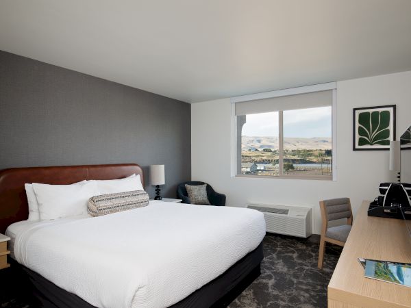 A modern hotel room with a bed, desk, chair, wall art, and a large window overlooking hills and a lake.