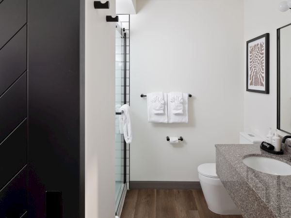 Modern bathroom with black door, glass shower, white towels, framed artwork, and a granite sink with toiletries.