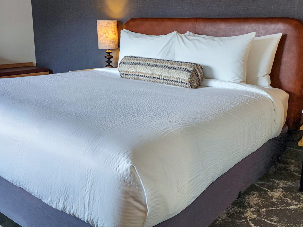 A neatly made bed with white linens, a brown headboard, two bedside lamps, and a decorative bolster pillow is shown in a room setting.