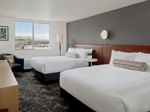 A modern hotel room with two white beds, a bedside lamp, and a large window showing mountains and a town outside.