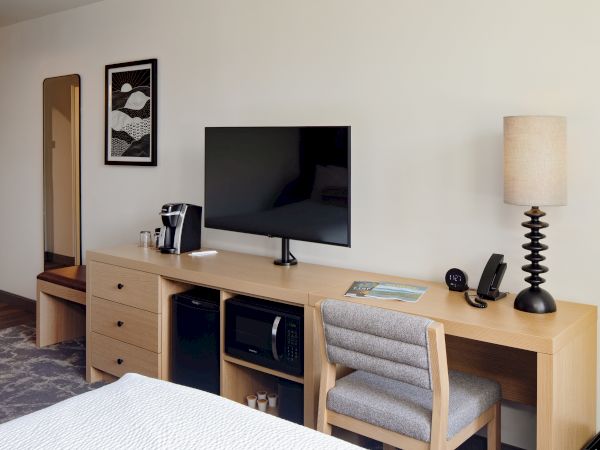 A modern hotel room with a flat-screen TV on a wooden console, coffee maker, alarm clock, and a bedside lamp.