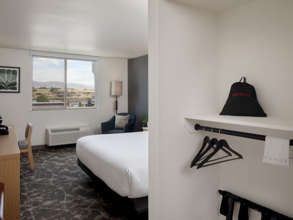 A hotel room with a bed, chair by a window, and a wall cubby holding a hair dryer and hangers.
