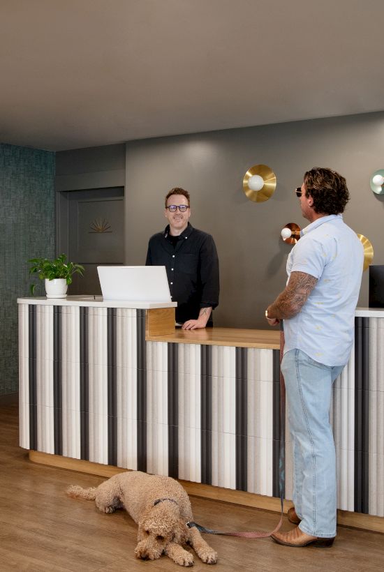 A hotel reception desk with two men chatting while a small dog walks in front of them.