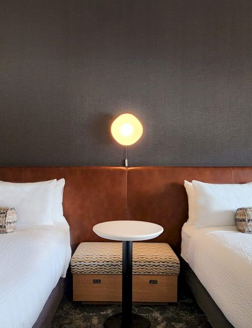 The image shows two beds with white linens, a central lamp on a table, and decorative pillows on each bed against a dark wall.