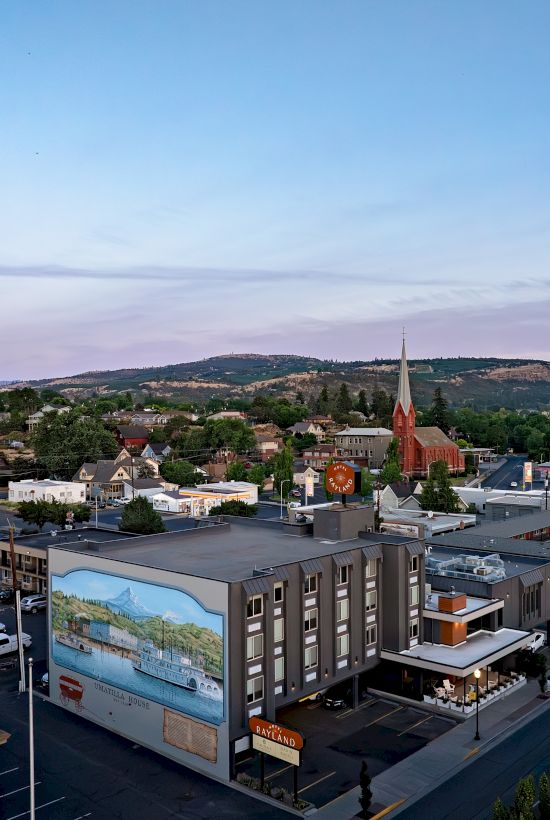 A small town view with a red church spire, hotels and streets, surrounded by green hills under a cloudy blue sky.