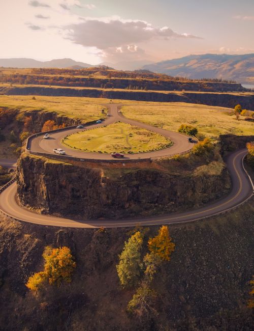 Aerial view of a winding two-lane road with cars in a grassy valley, surrounded by hills and mountains at sunset.