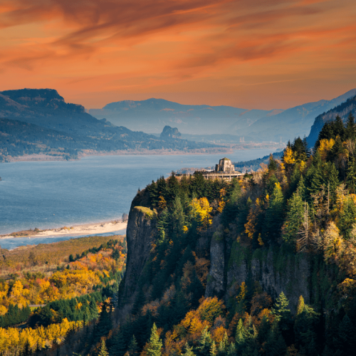 A scenic view of a river winding through a forested canyon, with a building perched on a cliff, under a vivid orange sky.