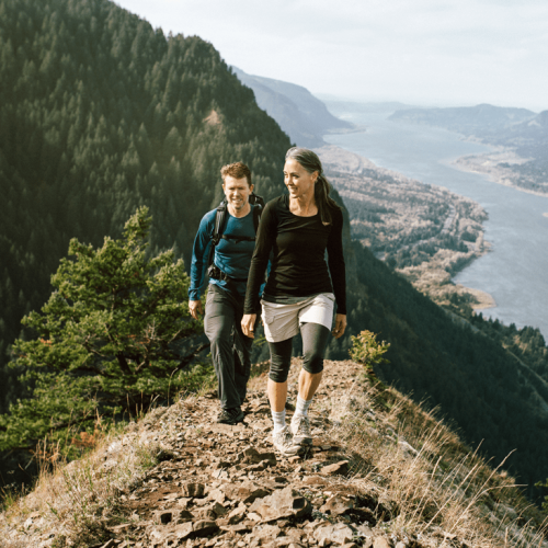 Two people are hiking on a rocky trail overlooking a river and forested mountains, enjoying the scenic view and outdoor adventure.