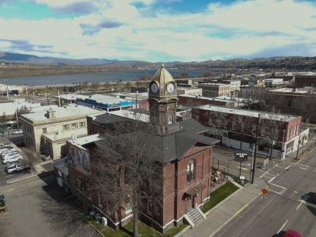 A historic clock tower in a small town overlooks a river and distant hills under a partly cloudy sky.