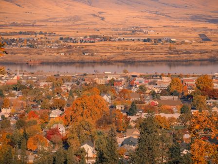 An autumn town stretches along a calm lake beneath rolling hills and fields at sunset, framed by trees with orange leaves.