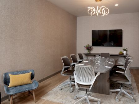 A modern conference room with a round table, swivel chairs, chandelier, wall TV, and small seating near the window.