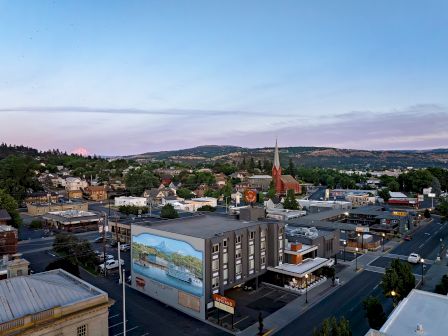 Aerial view of a small town at dusk with a red church steeple, hotels, roads, and rolling hills under a cloudy sky.