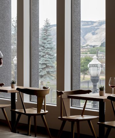 A man stands by a window in a modern caf&eacute;, holding a red wine glass with mountain scenery outside and patio-style tables set up beside him.