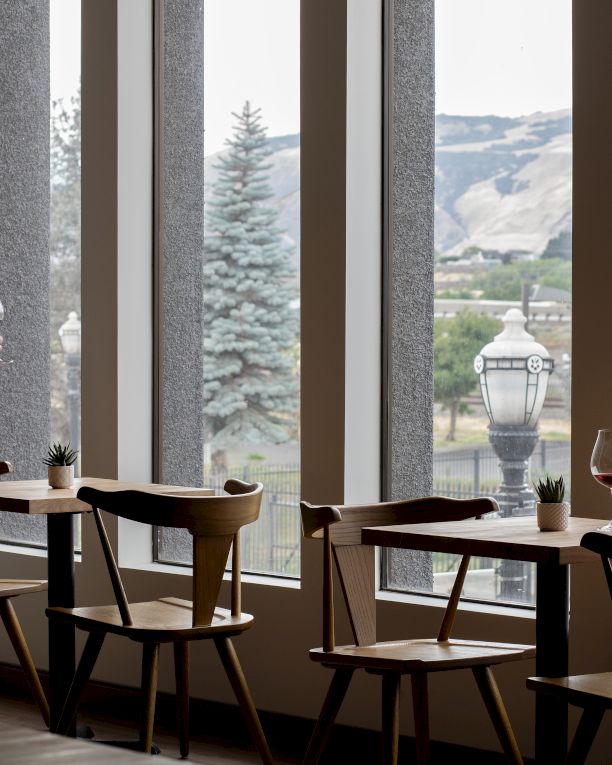 A man stands by a window in a modern caf&eacute;, holding a red wine glass with mountain scenery outside and patio-style tables set up beside him.
