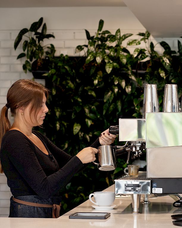 A barista makes espresso at a caf&eacute; machine next to a &ldquo;CAFE&hellip;COFFEEHOUSE&rdquo; logo.