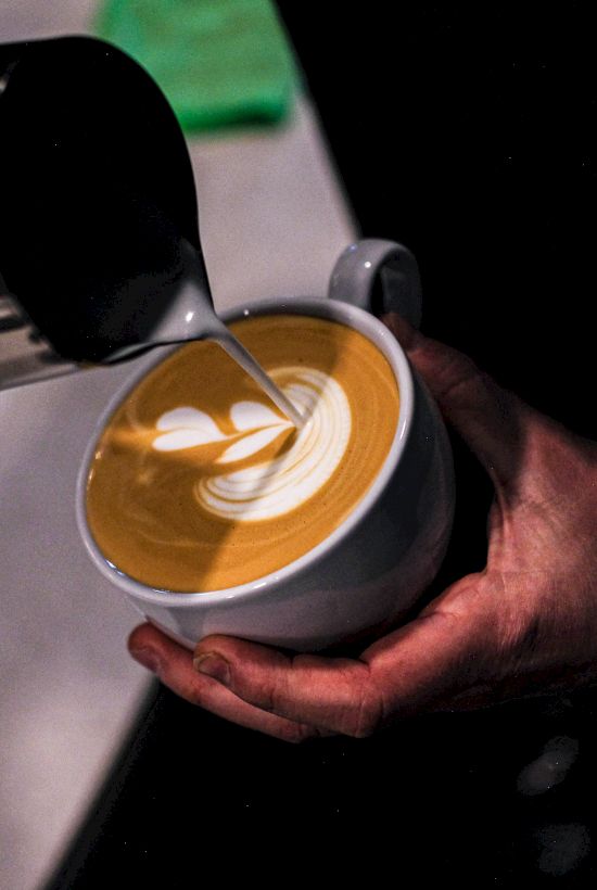 A barista pours steamed milk to create latte art in a cup, with a small heart/leaf pattern on the surface.