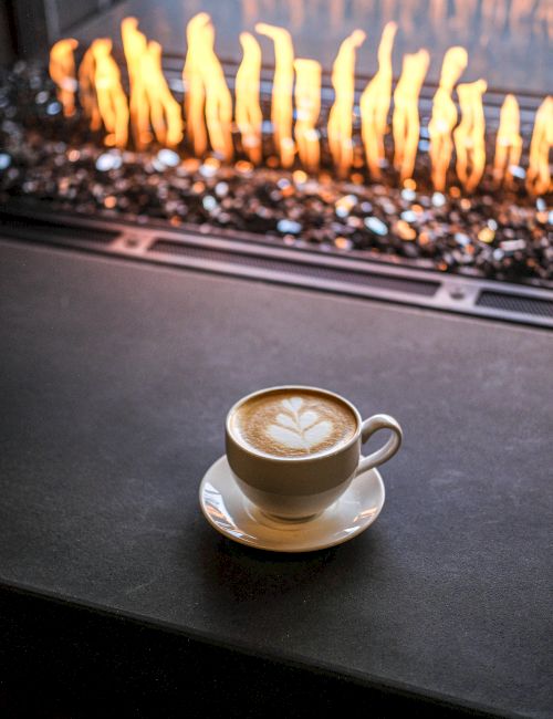 A cup of latte with latte art sits on a saucer in front of a fireplace with orange flames, cozy and warm.