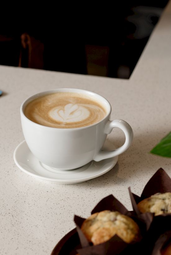 A cappuccino with leaf latte art sits on a table beside a chocolate pastry and a colorful placemat, in a cozy cafe.