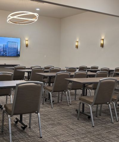 A conference or meeting room set up with rows of tables and chairs facing a wall-mounted screen.