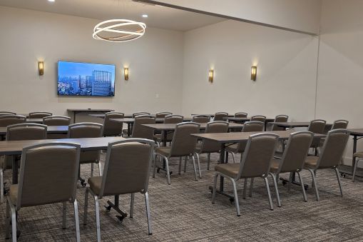 A conference or meeting room set up with rows of tables and chairs facing a wall-mounted screen.