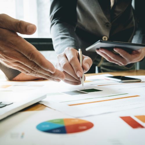 Two people are discussing documents and graphs on a table, with one holding a tablet and the other pointing and writing with a pen.