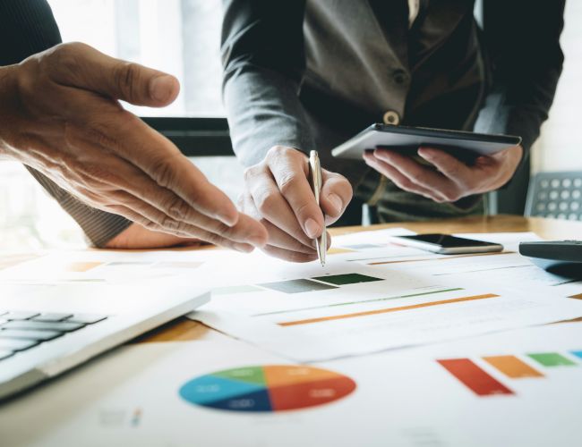 Two people are discussing documents and graphs on a table, with one holding a tablet and the other pointing and writing with a pen.