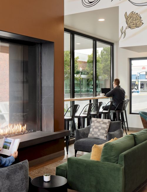 A modern lounge with a lit fireplace, comfy seating, people reading, and large windows opening to a bright street view.