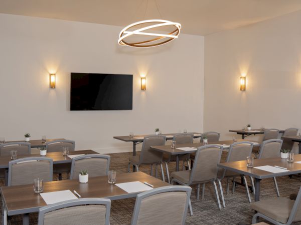 A modern meeting room with square tables, cushioned chairs, a wall TV, warm sconces, and a circular ceiling chandelier.