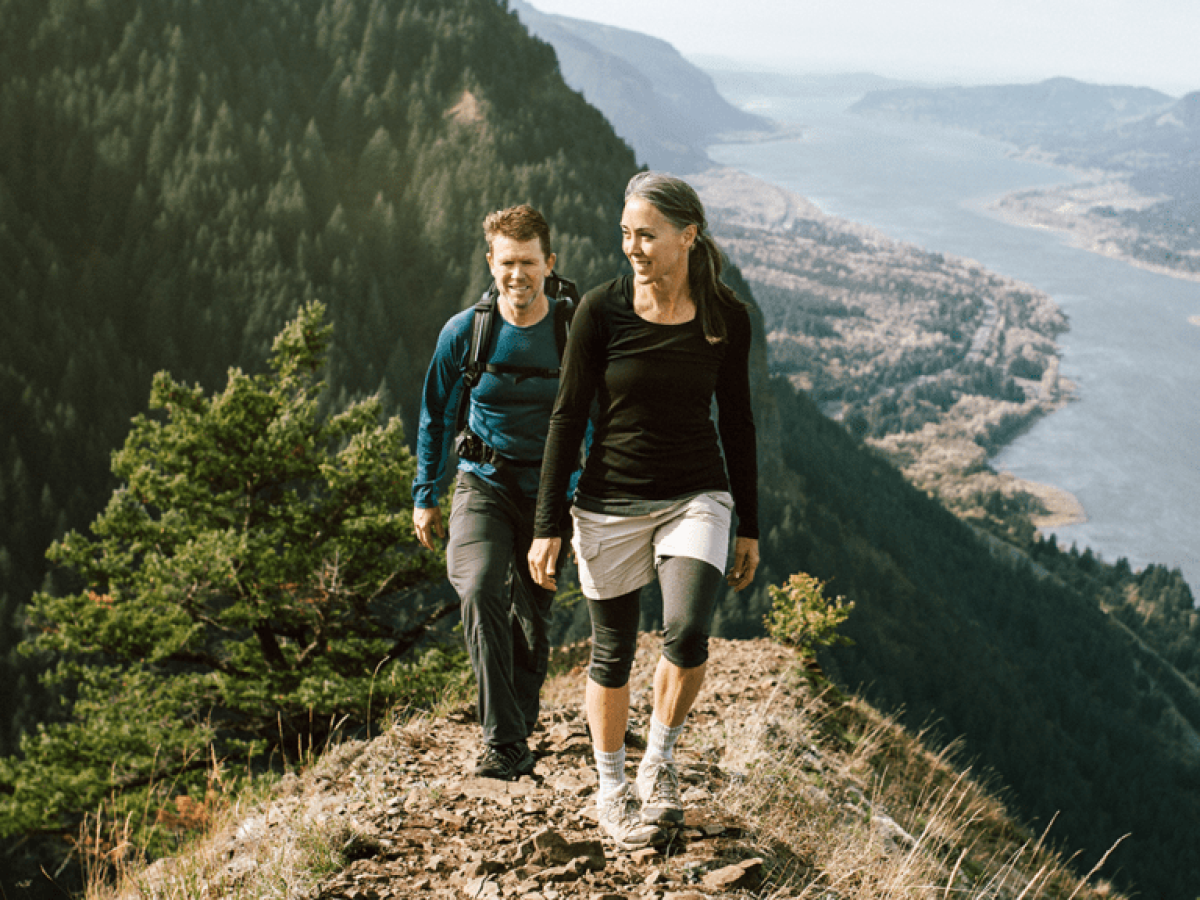 Two people hiking on a scenic mountain trail with a view of a river and dense forest in the background.