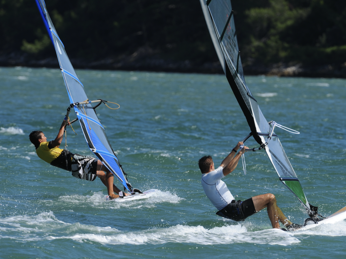 Two people are windsurfing on a sunny day, balancing on boards with sails over the water.