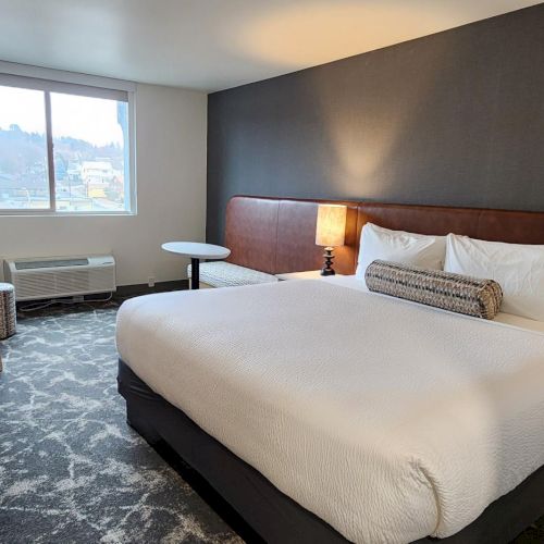 A modern hotel room with a large bed, two bedside lamps, a dark accent wall, a desk and chair, a TV, and a window letting in natural light.