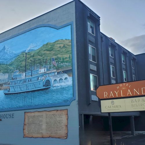 A tall glassy building with blue-tinted windows beside a smaller brick structure, city street in front, sunny day.