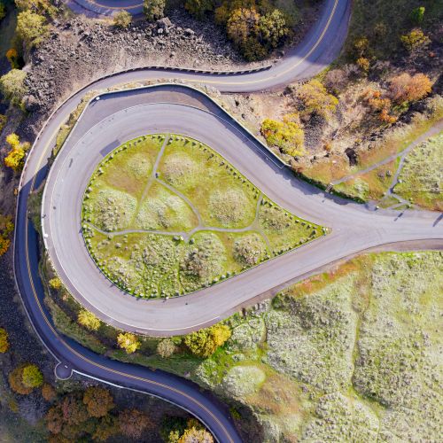 A winding road curves along a grassy hillside with guardrails, forming a loop around a small hilltop.