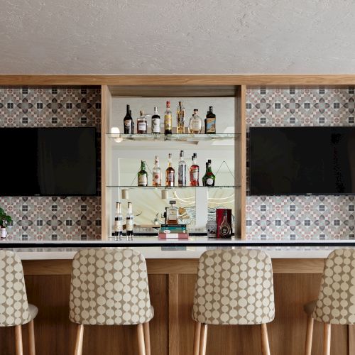 A built-in bar cabinet with mosaic tile, shelves of liquor bottles, two black screens, and three patterned stools beneath.