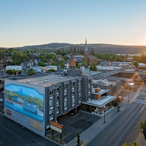 An aerial view of a small town at sunset, with a tall church steeple, roads, parked cars, and distant hills.