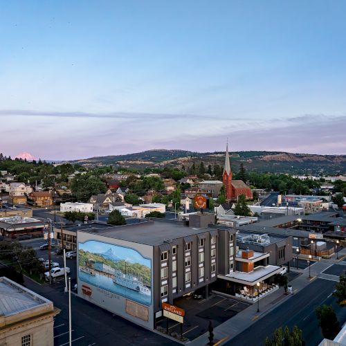A small town street scene with a red church steeple, hotels and shops in the foreground, and rolling hills under a cloudy sky.