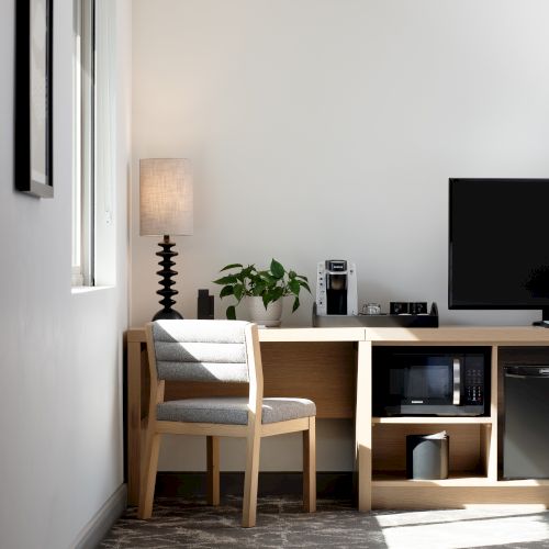 A tidy hotel office corner with a desk, flat-screen TV, coffee maker, lamp, potted plant, and a small chair.