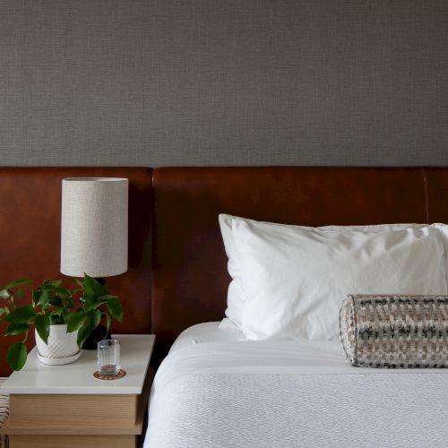 A hotel-style bed with pillows, a bedside table, a white lamp, and a small potted plant.