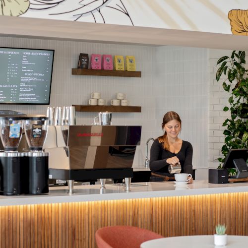 A caf&eacute; counter with a barista making coffee, an espresso machine, menu board, and indoor plants filling the wall.