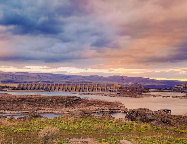 A scenic landscape with a river, a dam, rolling hills, and a dramatic sky during sunset or sunrise, giving a serene and vibrant atmosphere.