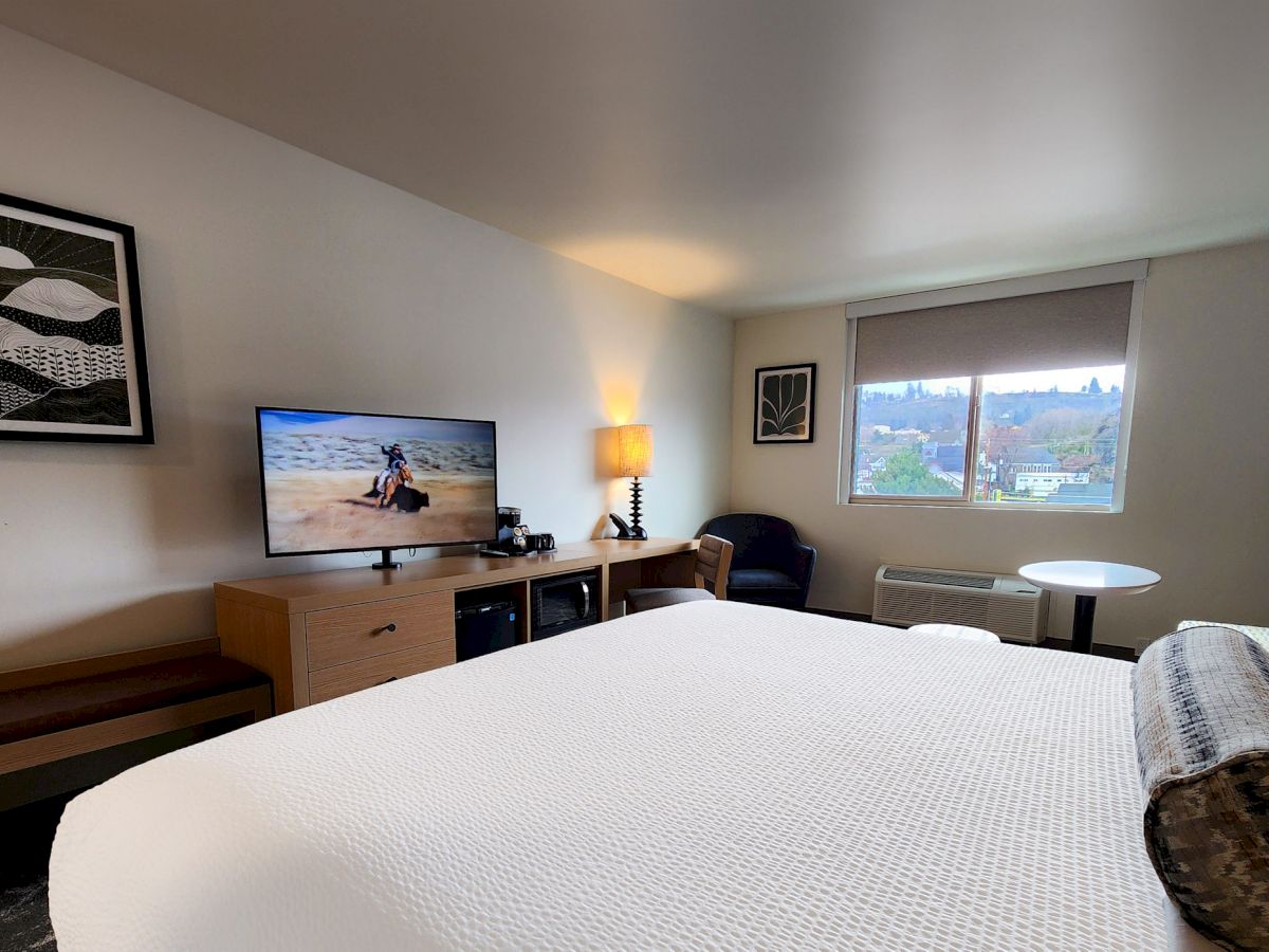 A modern hotel room with a large bed, wall art, TV, a window with a view, a small table, and a lamp in the corner by a chair.
