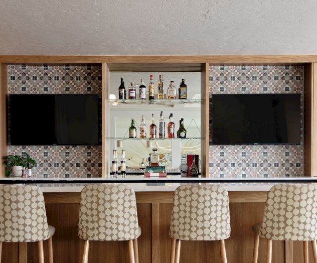 A built-in bar with mosaic tile, shelves of liquor bottles, two wall-mounted TVs, and three patterned stools in front.