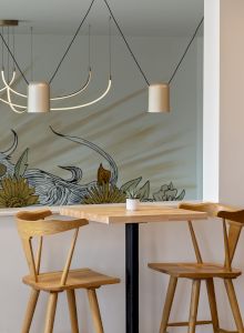 A bright caf&eacute; interior with geometric pendant lights, a floral mural wall, and wooden stools around a table.