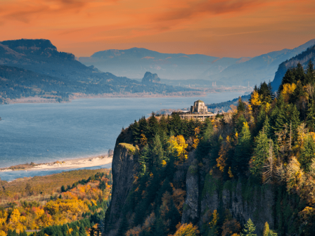 An autumn mountainside forest overlooks a calm blue lake at golden sunrise, with distant peaks fading into the mist.