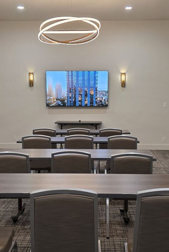 A conference room setup with multiple desks and chairs facing a wall-mounted screen; modern lighting and a circular ceiling light.