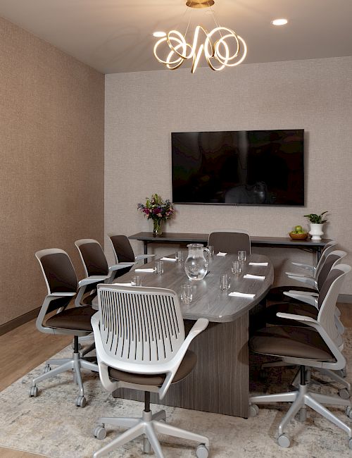 A modern office meeting room with a round glass table, chairs, TV, flowers, and a glowing circular chandelier.